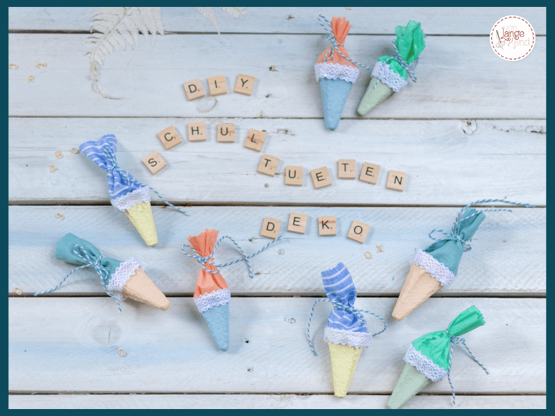 Selbst gebastelte Mini-Schultüten aus Eierkartons liegen zusammen mit Holzbuchstaben auf einem weißen Holzhintergrund.